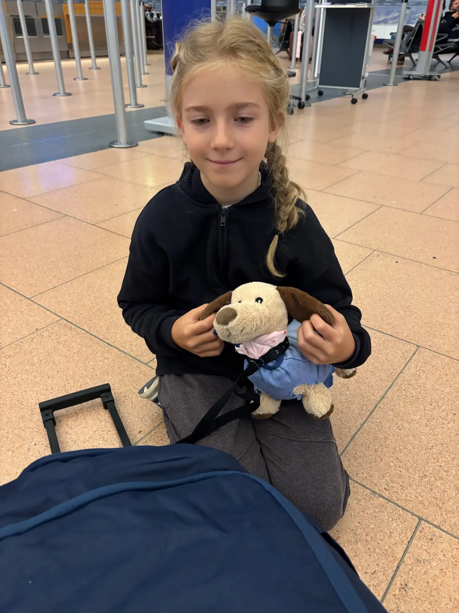 Merle sitzt am Flughafen auf ihrem Koffer, mit einem Kuscheltier